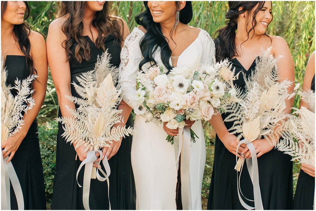 bride holding white bouquet with anemones, bridesmaids wearing black dresses and holding dried pampas grass bouquets